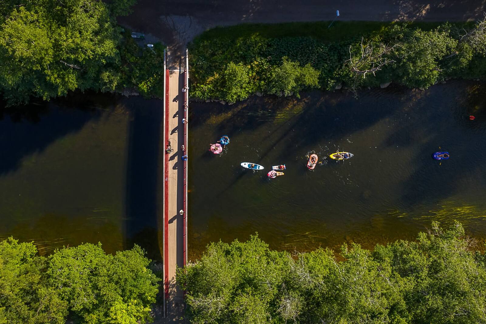 Flygbild över den röda gångbron där floatare, SUP:ar och kajaker passerar under genom grön skog